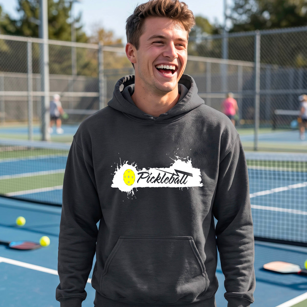 A smiling man wearing a dark gray hoodie with a 'Pickleball' splatter graphic on the chest stands on an outdoor pickleball court. 