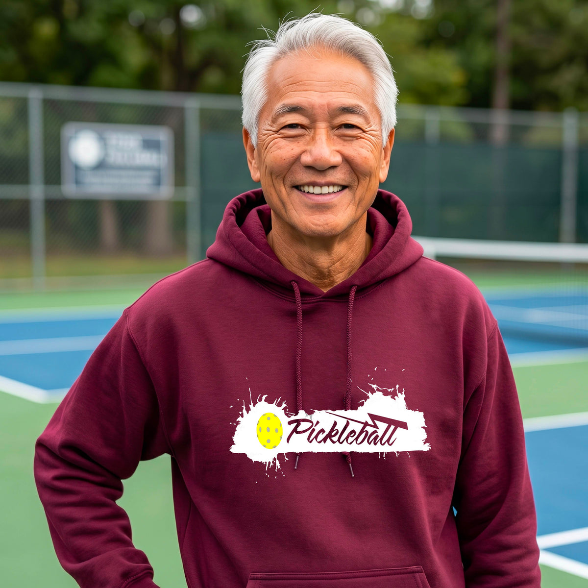 A smiling older man wearing a maroon hoodie with a 'Pickleball' splatter graphic on the chest stands on an outdoor pickleball court. 