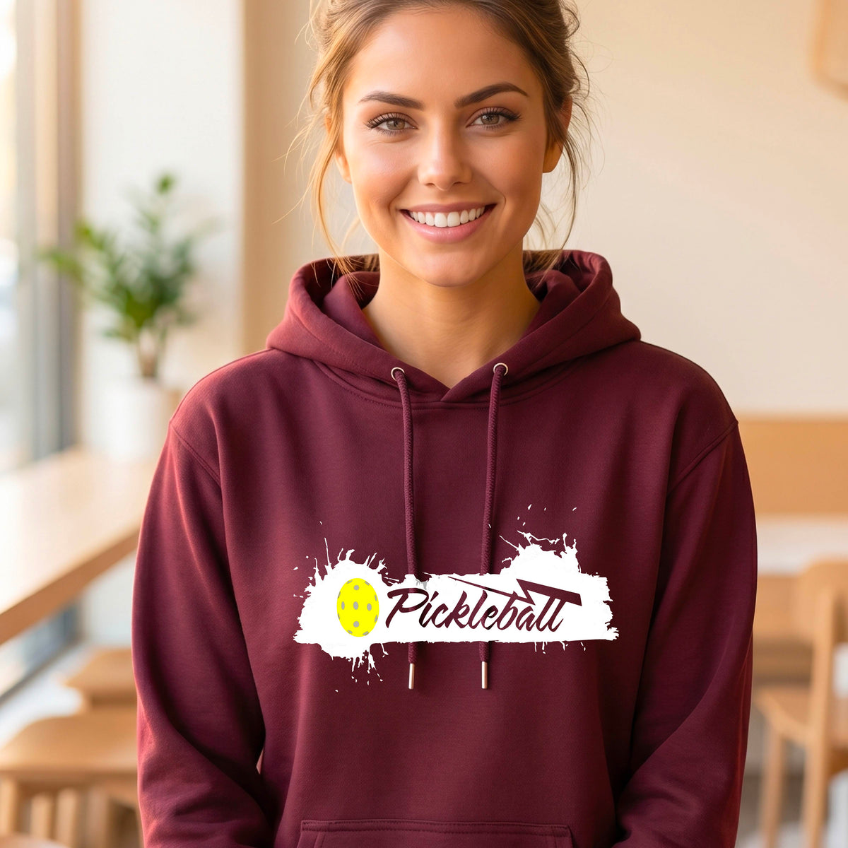 A smiling woman wearing a maroon hoodie with a 'Pickleball' splatter graphic on the chest stands on an outdoor pickleball court. 
