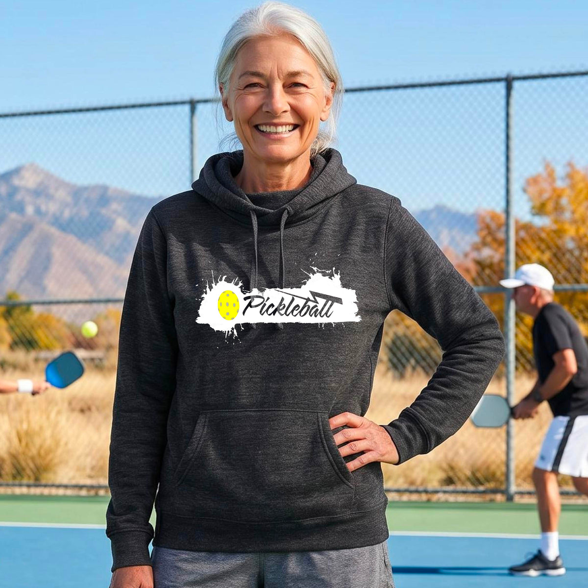 A smiling older woman stands on an outdoor pickleball court wearing a blackl heather hoodie with a white and yellow 'Pickleball' splatter graphic on the chest. 