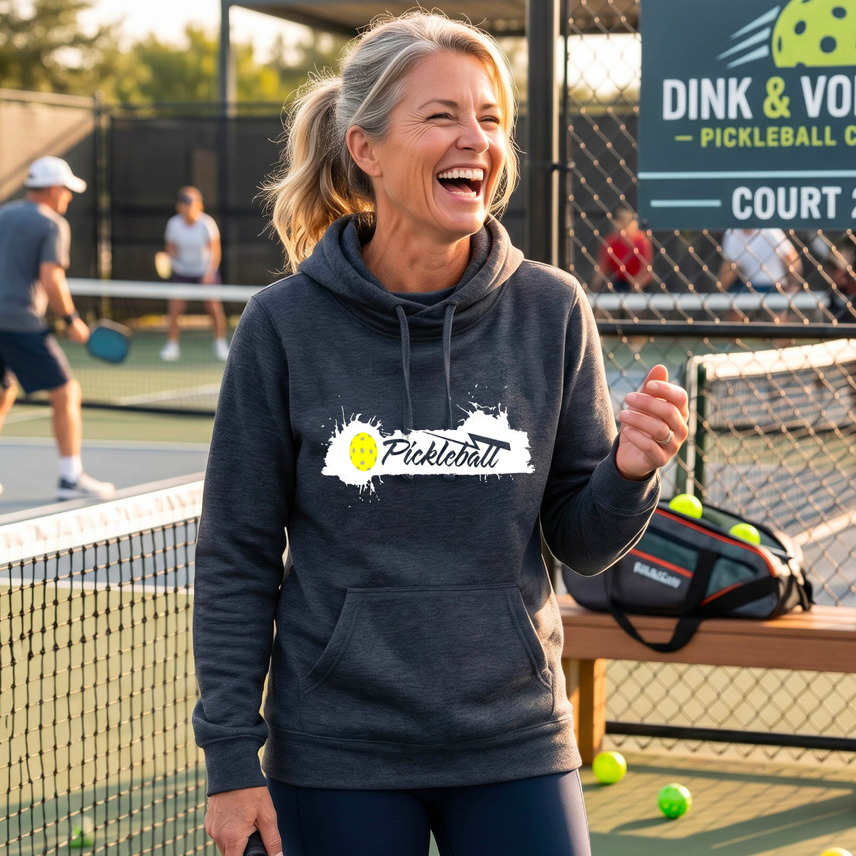 A smiling woman stands on an outdoor pickleball court wearing a navy blue hoodie with a white and yellow 'Pickleball' splatter graphic on the chest. 
