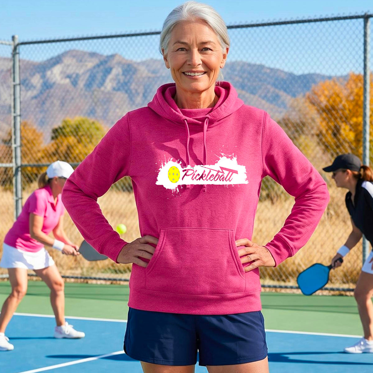 A smiling woman stands on an outdoor pickleball court wearing a pink hoodie with a white and yellow 'Pickleball' splatter graphic on the chest. 