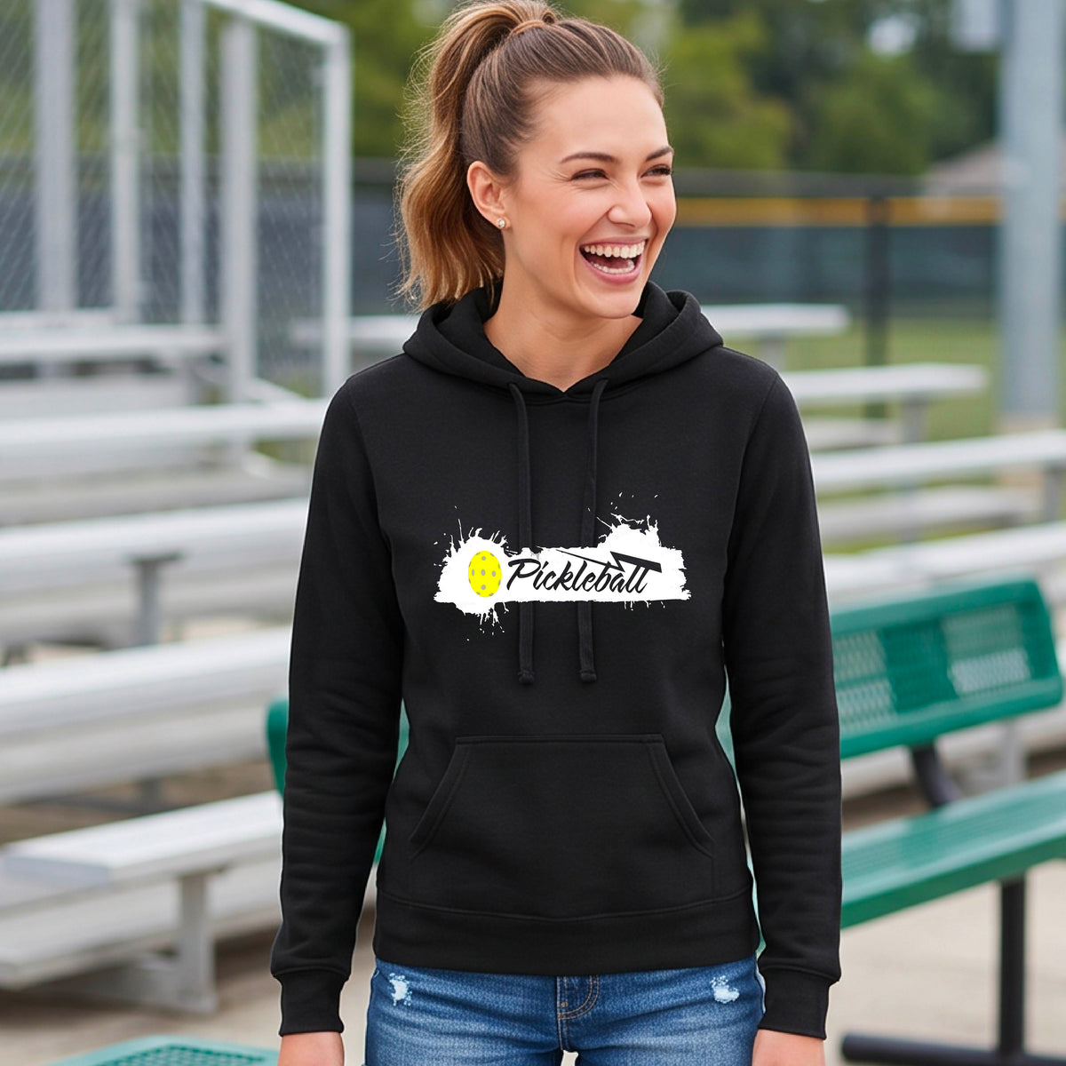 A woman wearing a black fitted hoodie with a 'Pickleball' graphic on the chest stands on a pickleball court. The graphic features a white paint splatter background with a yellow pickleball.