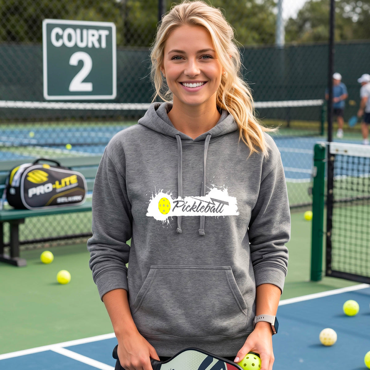 A woman wearing a gray fitted hoodie with a 'Pickleball' graphic on the chest stands on a pickleball court. The graphic features a white paint splatter background with a yellow pickleball.