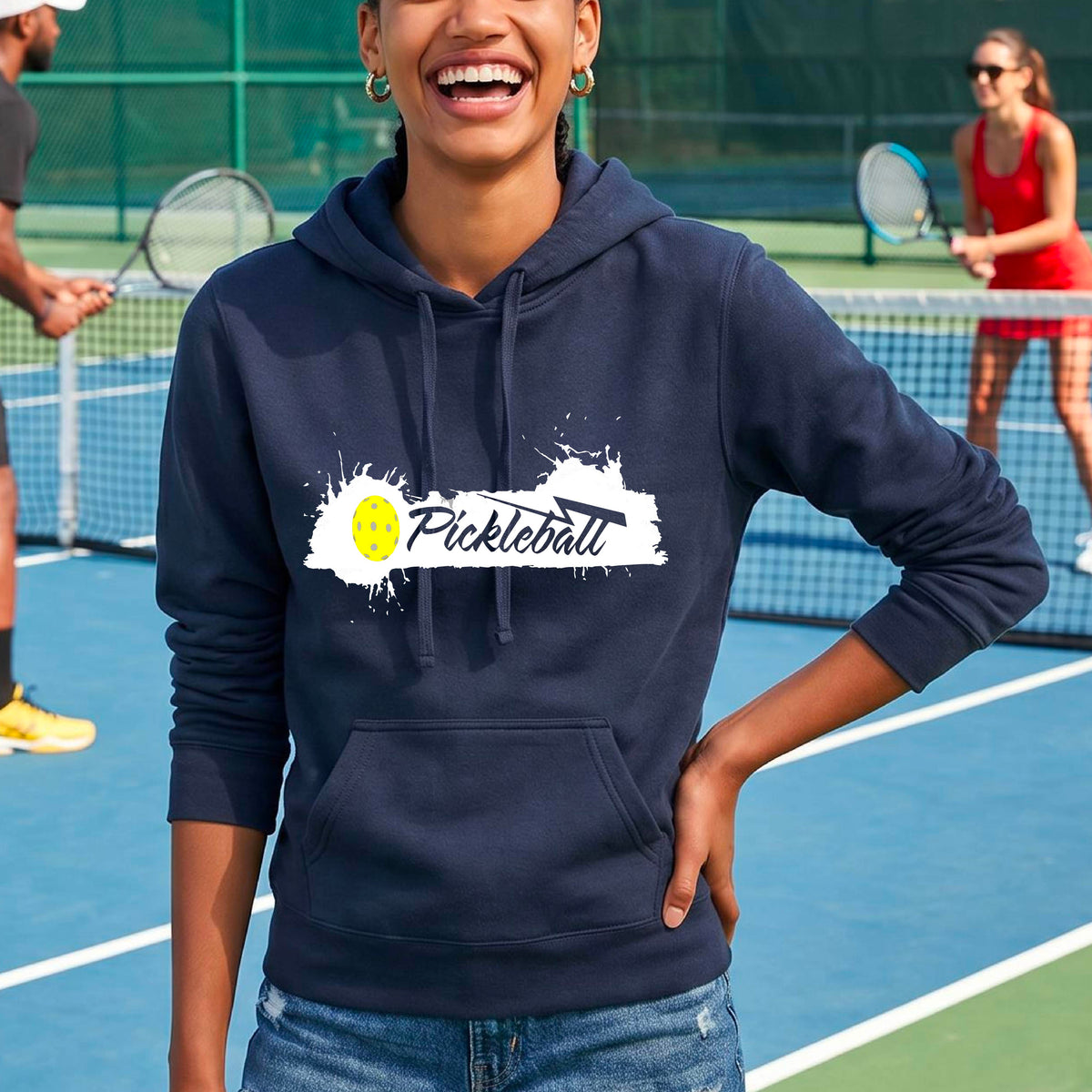 A woman wearing a navy blue fitted hoodie with a 'Pickleball' graphic on the chest stands on a pickleball court. The graphic features a white paint splatter background with a yellow pickleball.