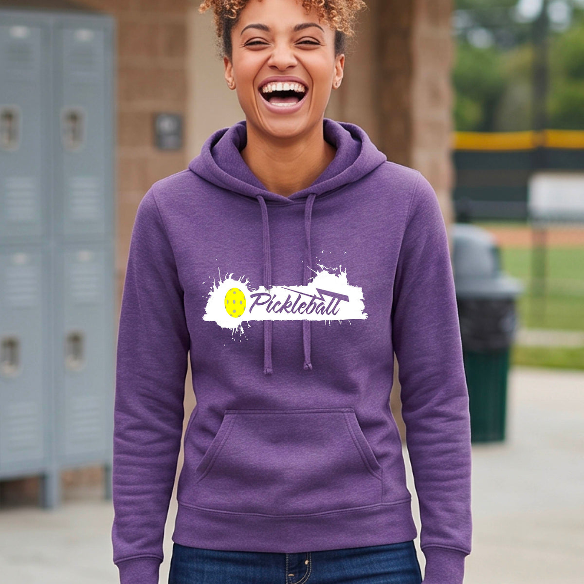 A woman wearing a purple fitted hoodie with a 'Pickleball' graphic on the chest. The graphic features a white paint splatter background with a yellow pickleball.