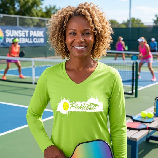 A smiling woman on an outdoor pickleball court wearing a bright lime green long-sleeve shirt with a white and yellow 'Pickleball' and splatrer graphic on the chest.