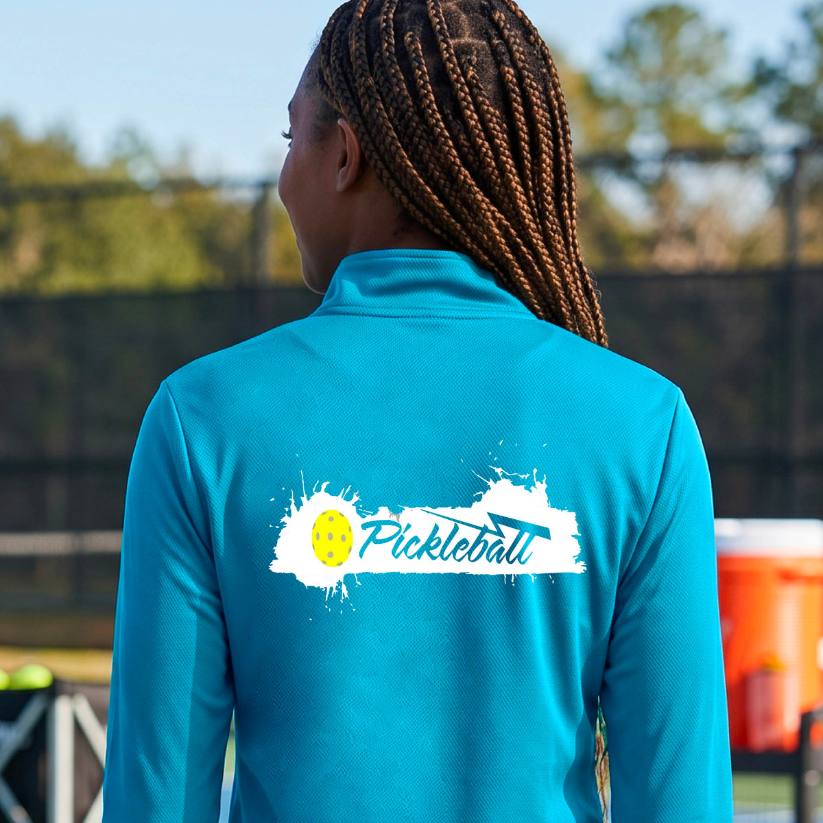 A woman viewed from behind wearing a bright blue long-sleeve quarter-zip athletic top with a white and yellow 'Pickleball' splatter graphic on the back, standing on an outdoor pickleball court.