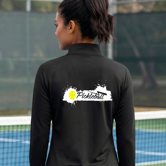 A woman viewed from behind wearing a black long-sleeve quarter-zip athletic top with a white and yellow 'Pickleball' splatter graphic on the back, standing on an outdoor pickleball court.