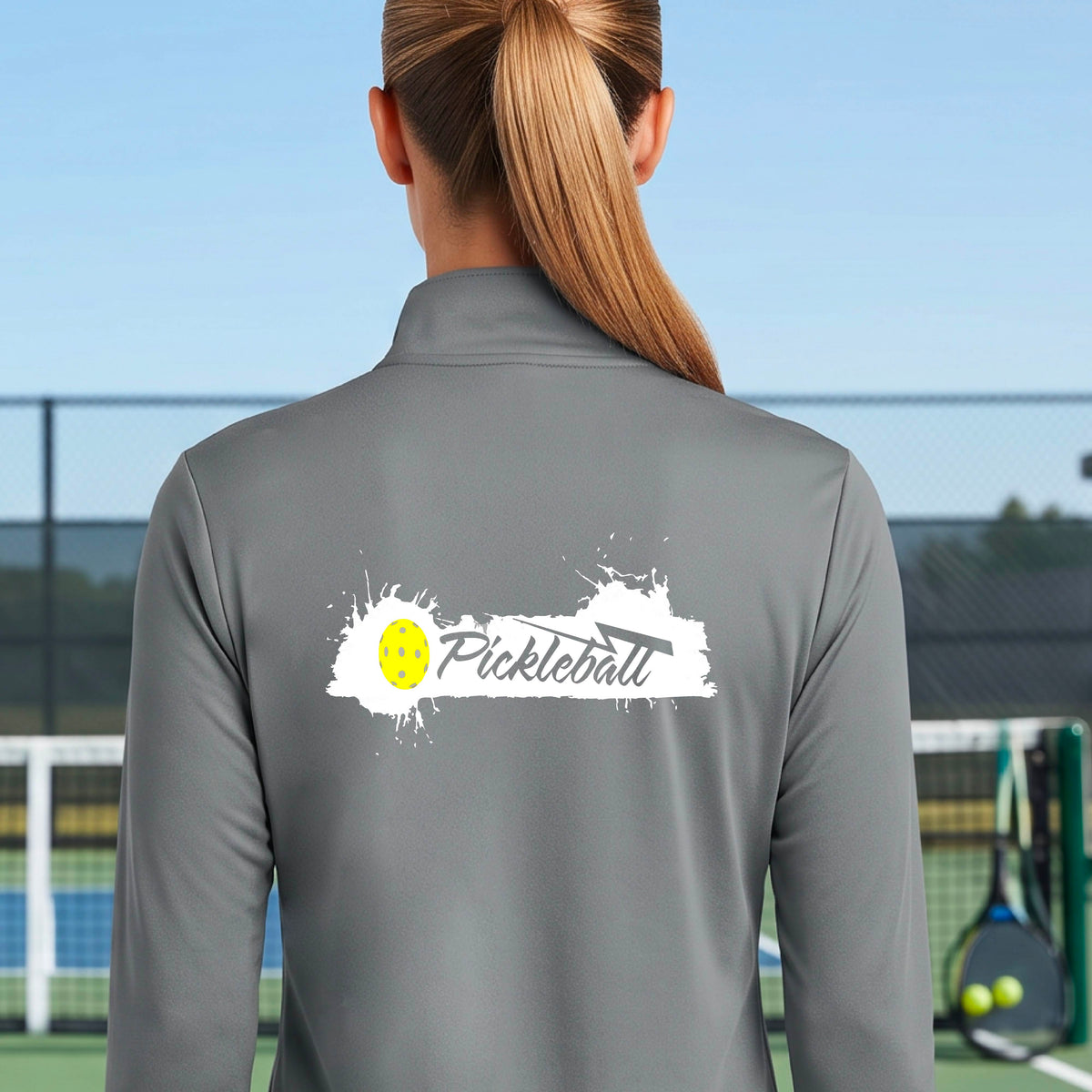 A woman viewed from behind wearing a gray long-sleeve quarter-zip athletic top with a white and yellow 'Pickleball' splatter graphic on the back, standing on an outdoor pickleball court.