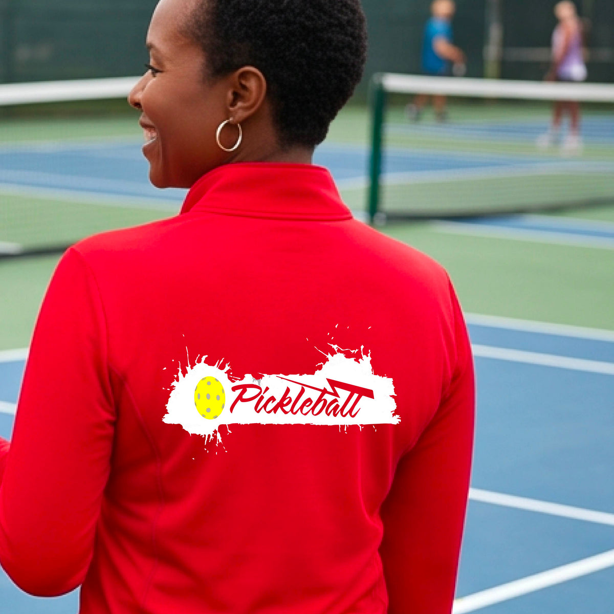 A woman viewed from behind wearing a red long-sleeve quarter-zip athletic top with a white and yellow 'Pickleball' splatter graphic on the back, standing on an outdoor pickleball court.
