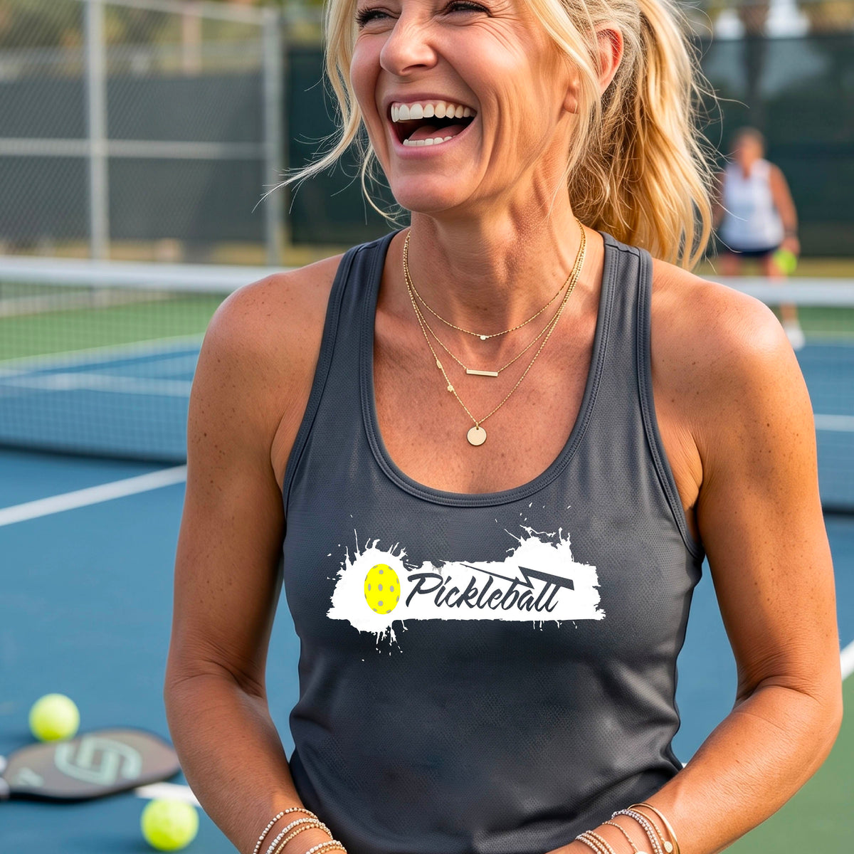 A smiling woman on a pickleball court wearing a daak gray racerback athletic tank top with a white and yellow 'Pickleball' graphic on the front.