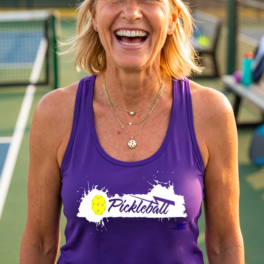 A smiling woman on a pickleball court wearing a purple racerback athletic tank top with a white and yellow 'Pickleball' graphic on the front.