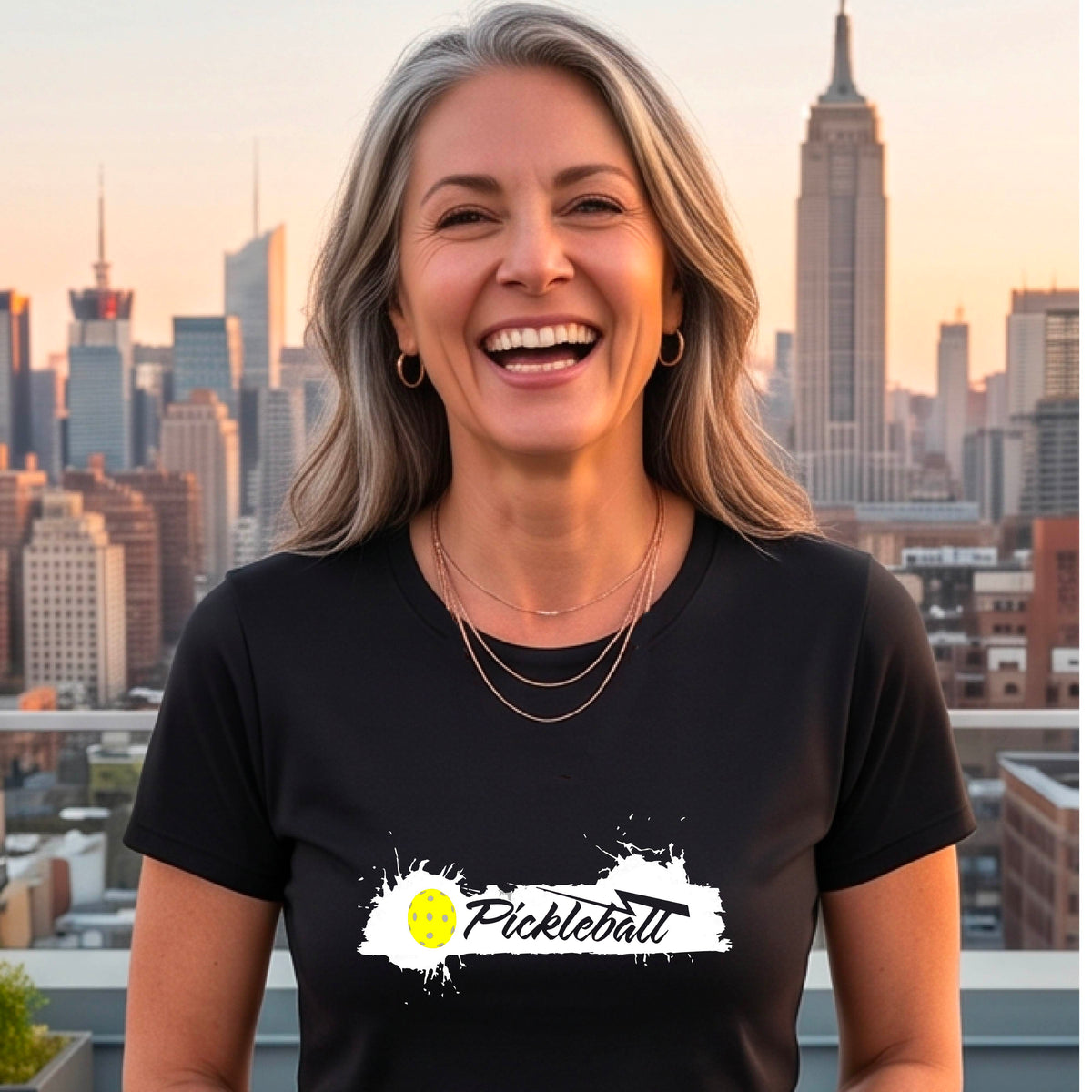 A woman wearing a black short-sleeve t-shirt top with a white paint splatter graphic that reads 'Pickleball' in a fun script. with a yellow pickleball.