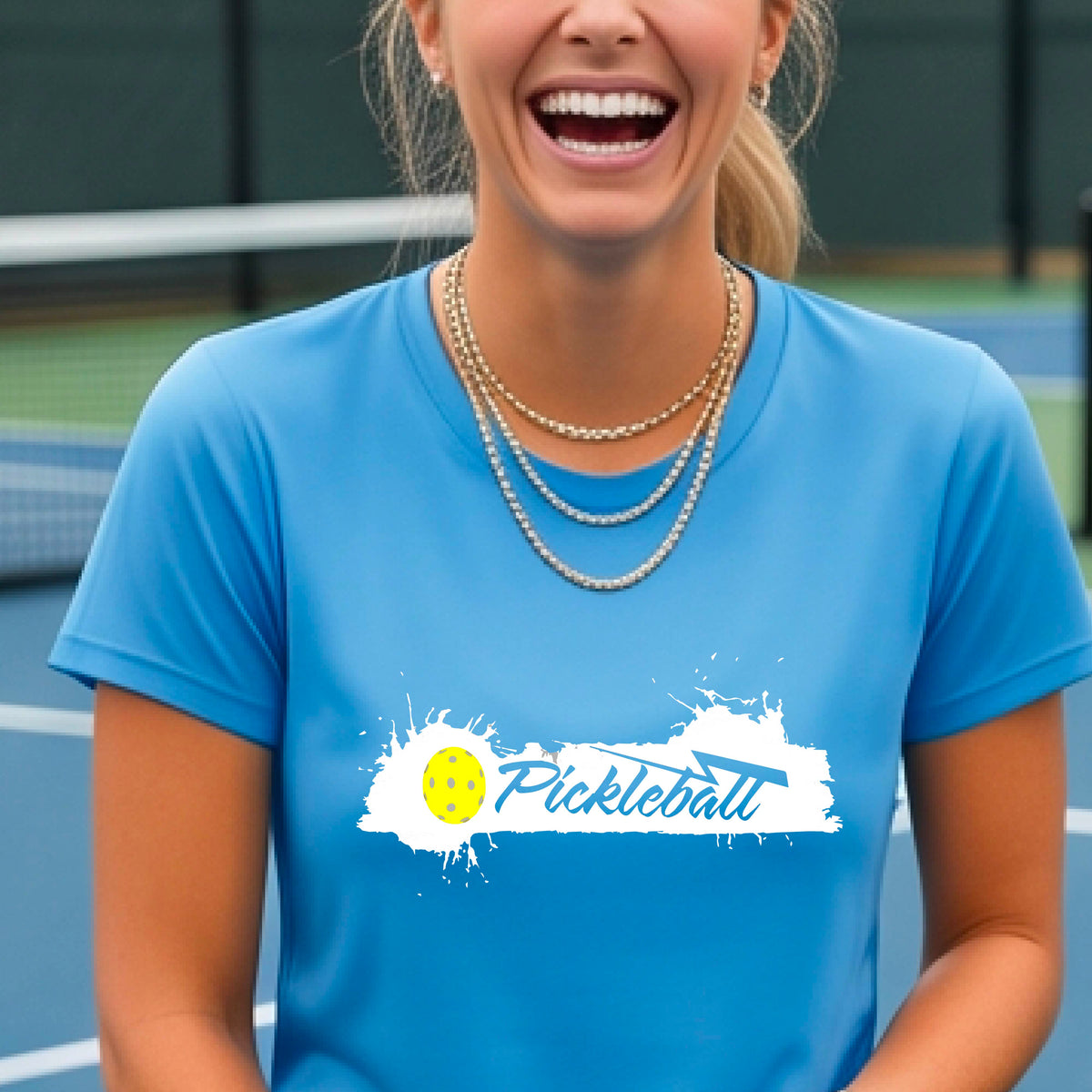 A woman wearing a light blue short-sleeve t-shirt top with a white paint splatter graphic that reads 'Pickleball' in a fun script. with a yellow pickleball.