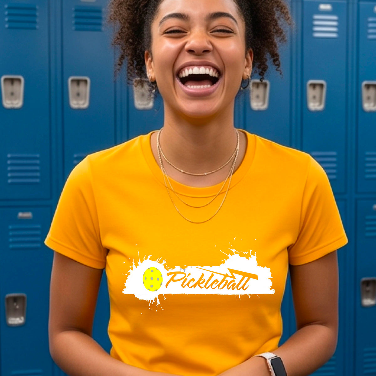 A woman wearing a mustard yellow gold  short-sleeve t-shirt top with a white paint splatter graphic that reads 'Pickleball' in a fun script. with a yellow pickleball.
