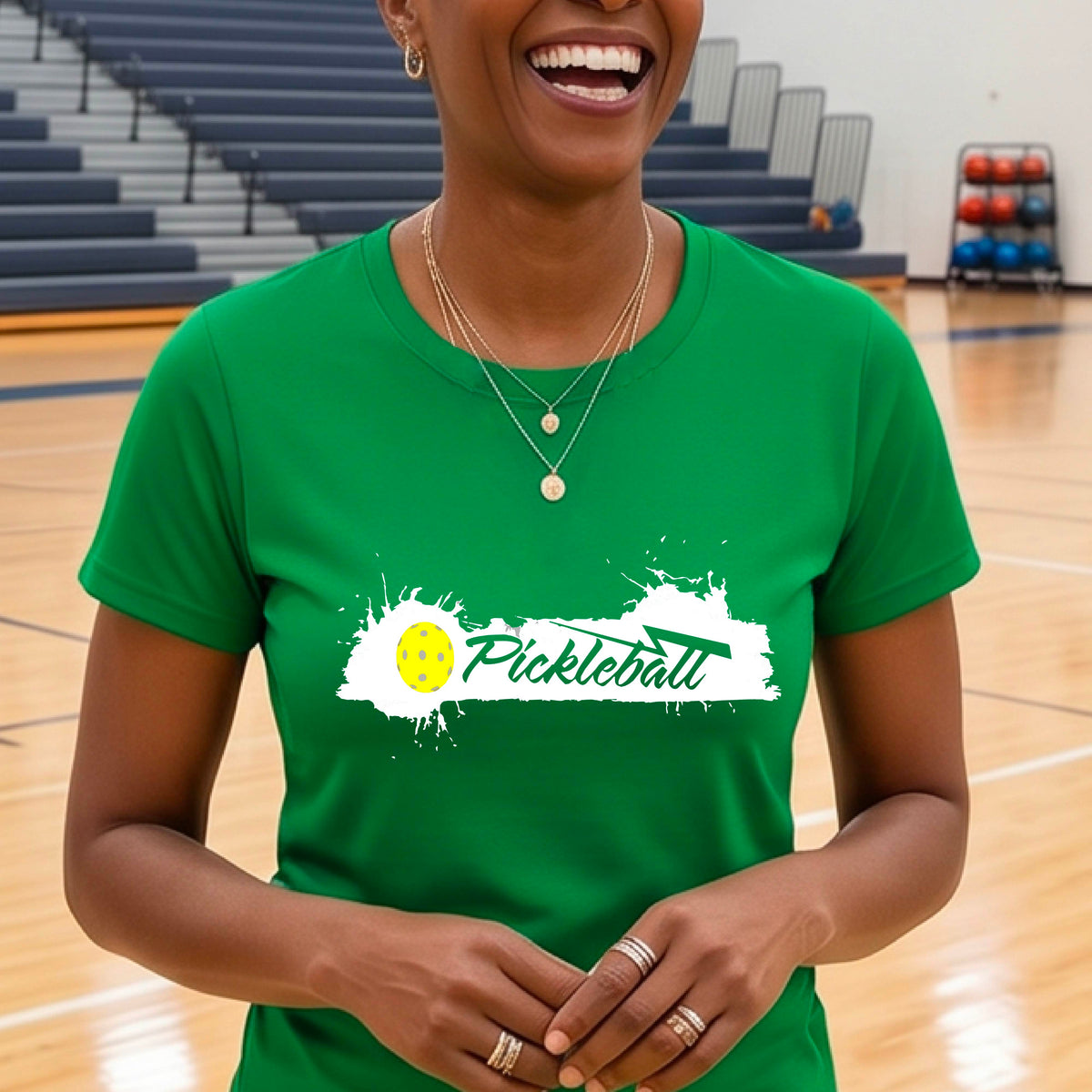 A woman wearing a green short-sleeve t-shirt top with a white paint splatter graphic that reads 'Pickleball' in a fun script. with a yellow pickleball.