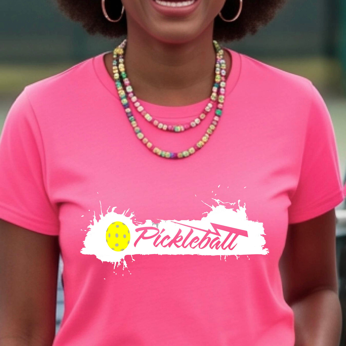 A woman wearing a pink short-sleeve t-shirt top with a white paint splatter graphic that reads 'Pickleball' in a fun script. with a yellow pickleball.