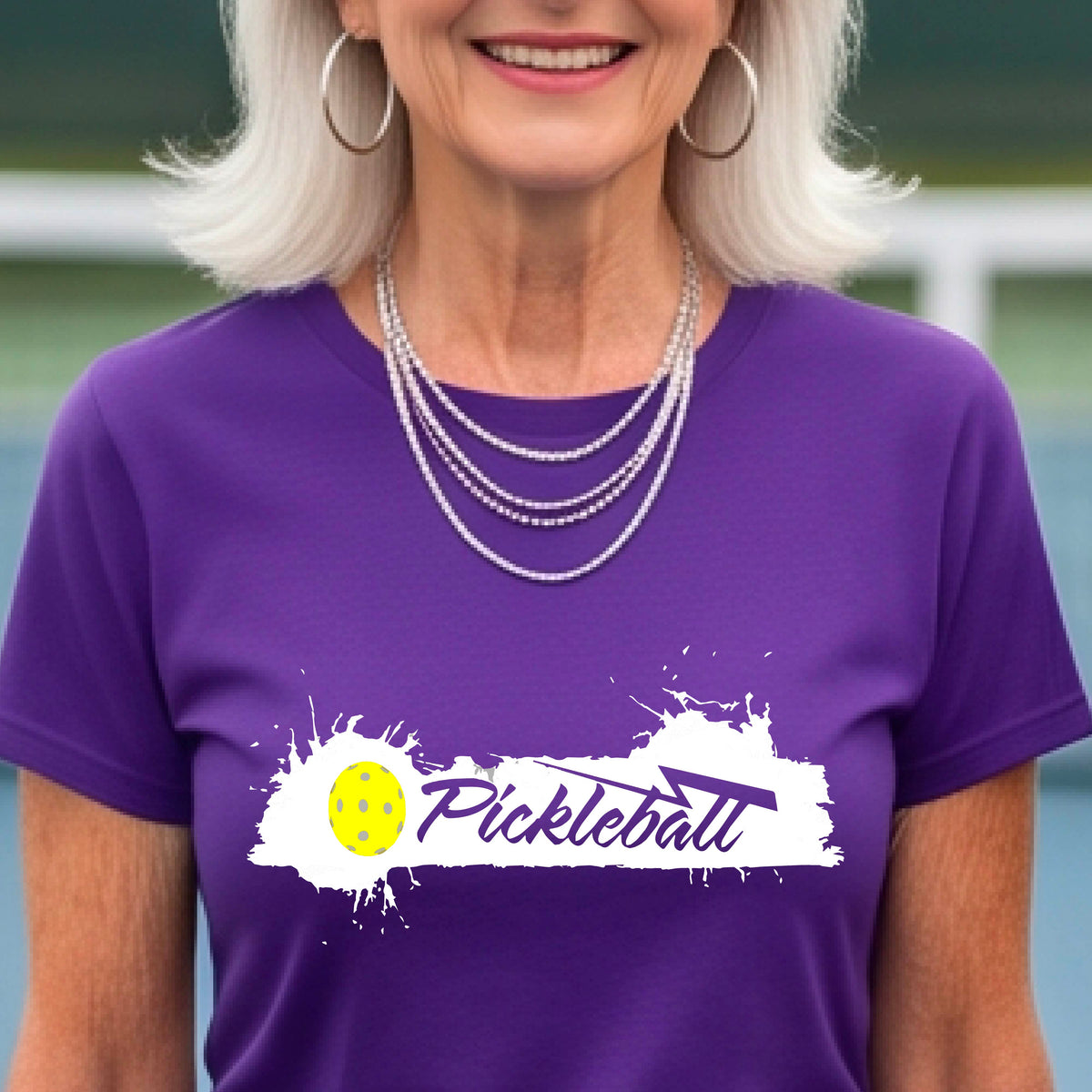 A woman wearing a purple short-sleeve t-shirt top with a white paint splatter graphic that reads 'Pickleball' in a fun script. with a yellow pickleball.