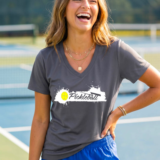 A woman wearing a dark gray V-neck t-shirt with a white and yellow 'Pickleball' splatter graphic on the front..