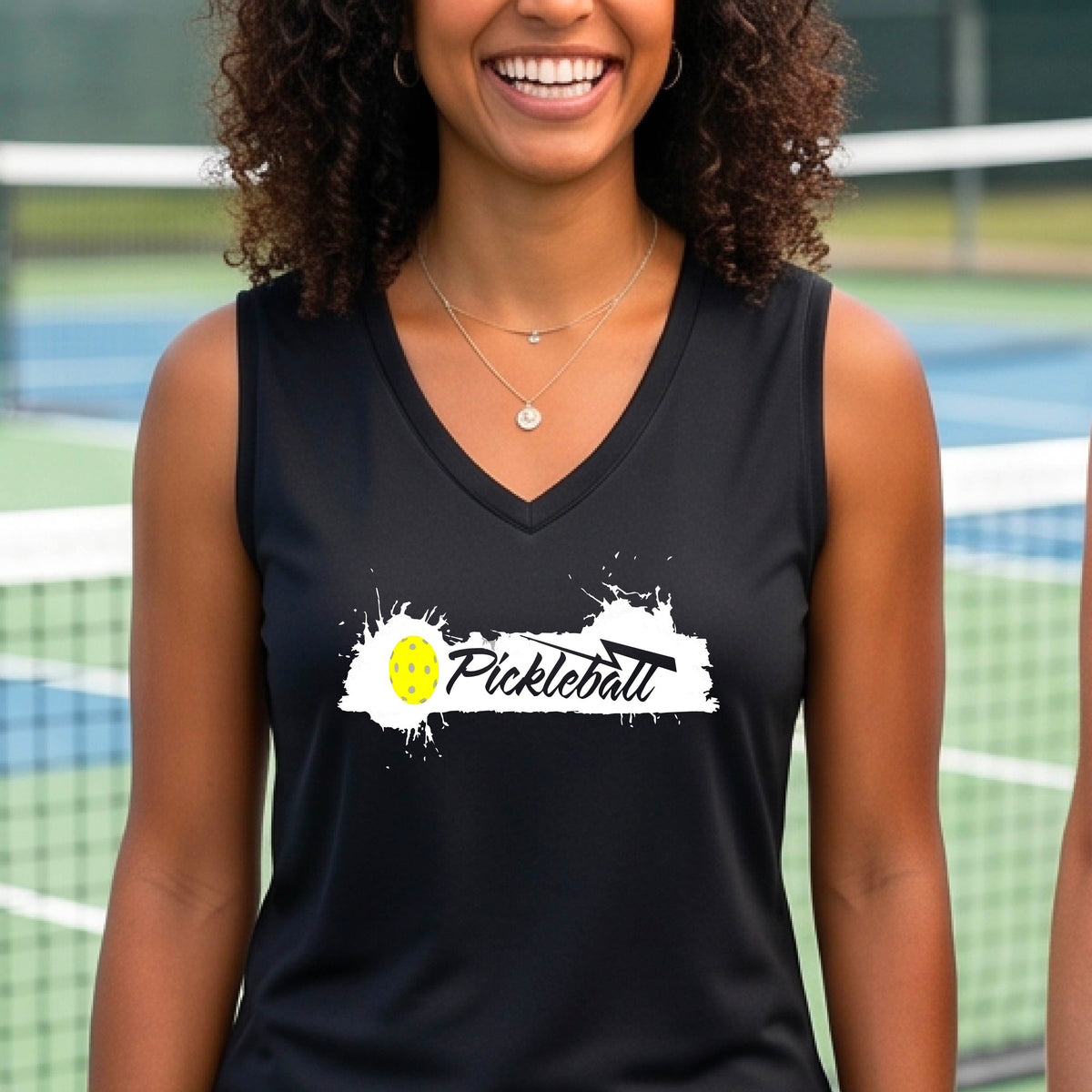 A smiling woman wearing a black sleeveless V-neck athletic top astanding on a pickleball court. The top has a white paint-splatter graphic with a yellow pickleball and the word 'Pickleball' across the chest.