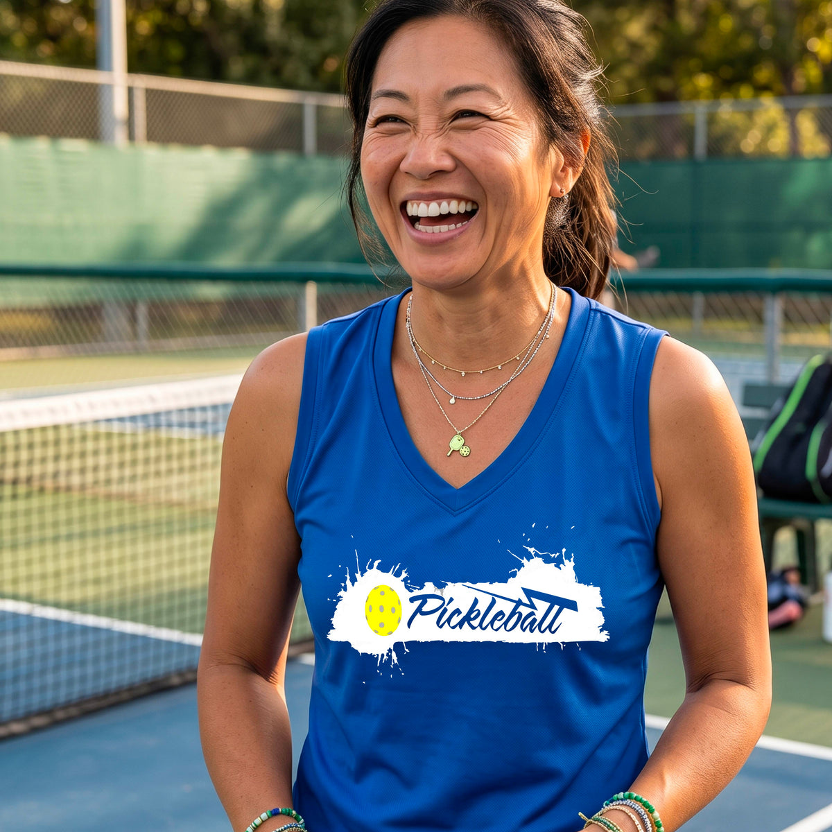 A smiling woman wearing a royal blue sleeveless V-neck athletic top astanding on a pickleball court. The top has a white paint-splatter graphic with a yellow pickleball and the word 'Pickleball' across the chest.