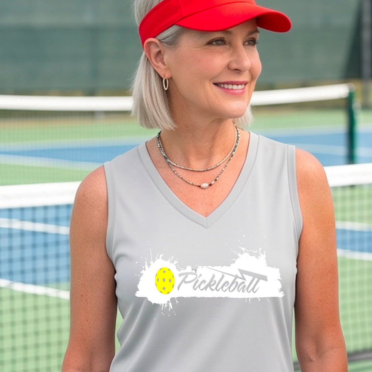 A smiling woman wearing a silver gray sleeveless V-neck athletic top astanding on a pickleball court. The top has a white paint-splatter graphic with a yellow pickleball and the word 'Pickleball' across the chest.