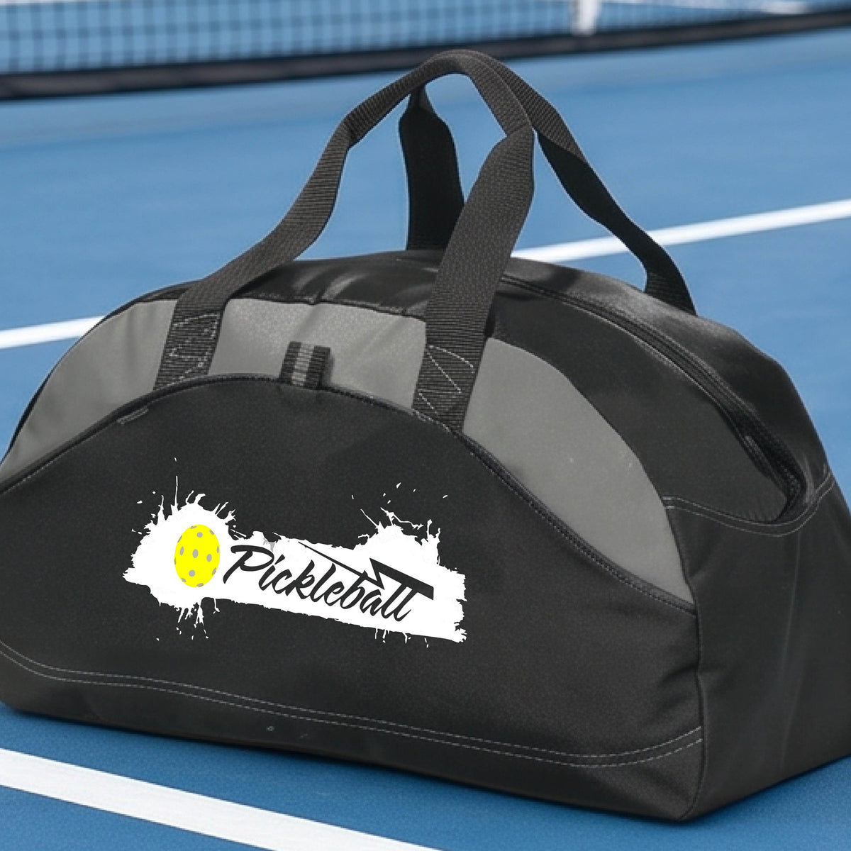A black and gray pickleball duffel bag with a white and yellow 'Pickleball' splatter graphic, resting on a pickleball court.