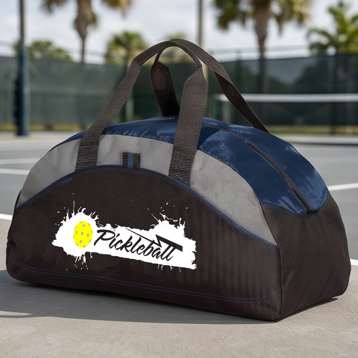 A navy blue, black and gray pickleball duffel bag with a white and yellow 'Pickleball' splatter graphic, resting on a pickleball court.