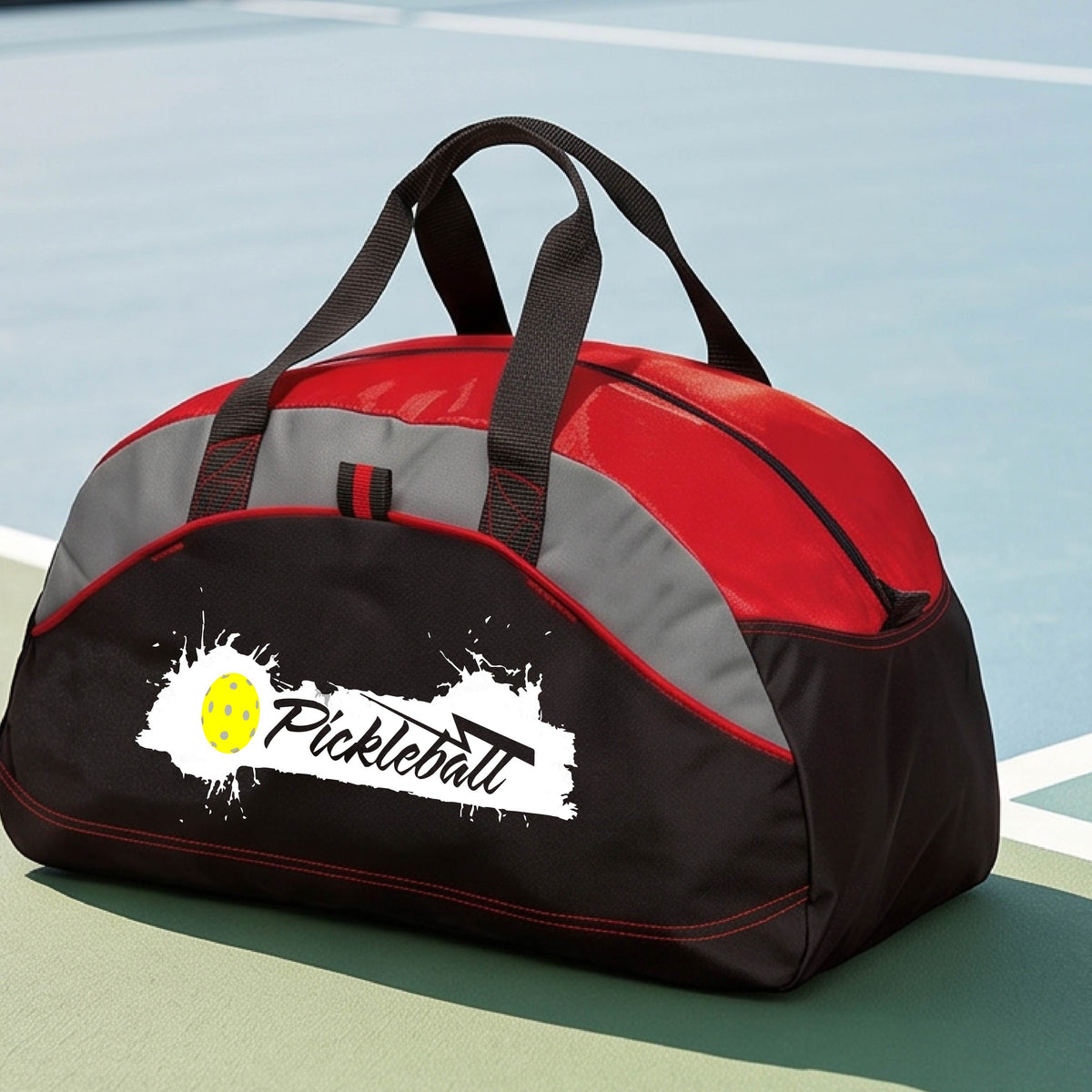A red and gray pickleball duffel bag with a white and yellow 'Pickleball' splatter graphic, resting on a pickleball court.