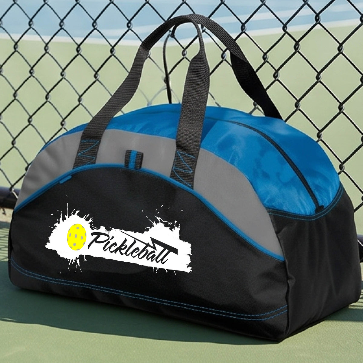 A royal blue, black and gray pickleball duffel bag with a white and yellow 'Pickleball' splatter graphic, resting on a pickleball court.