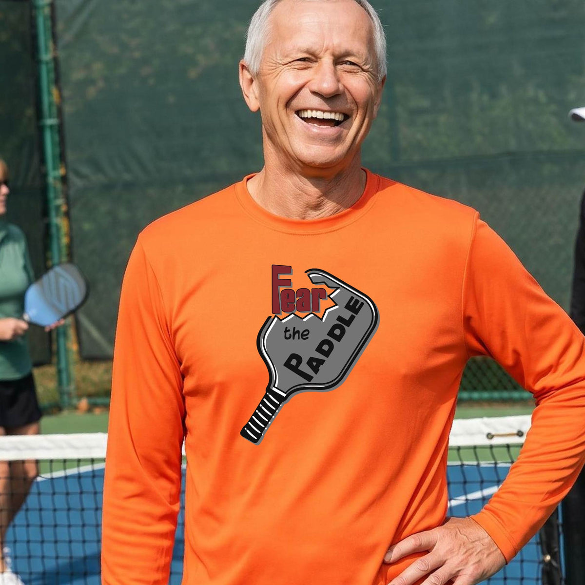 A man wearing a long-sleeve orange shirt with a pickleball graphic of the words 'FEAR THE PADDLE' cut into a pickleball paddle. 