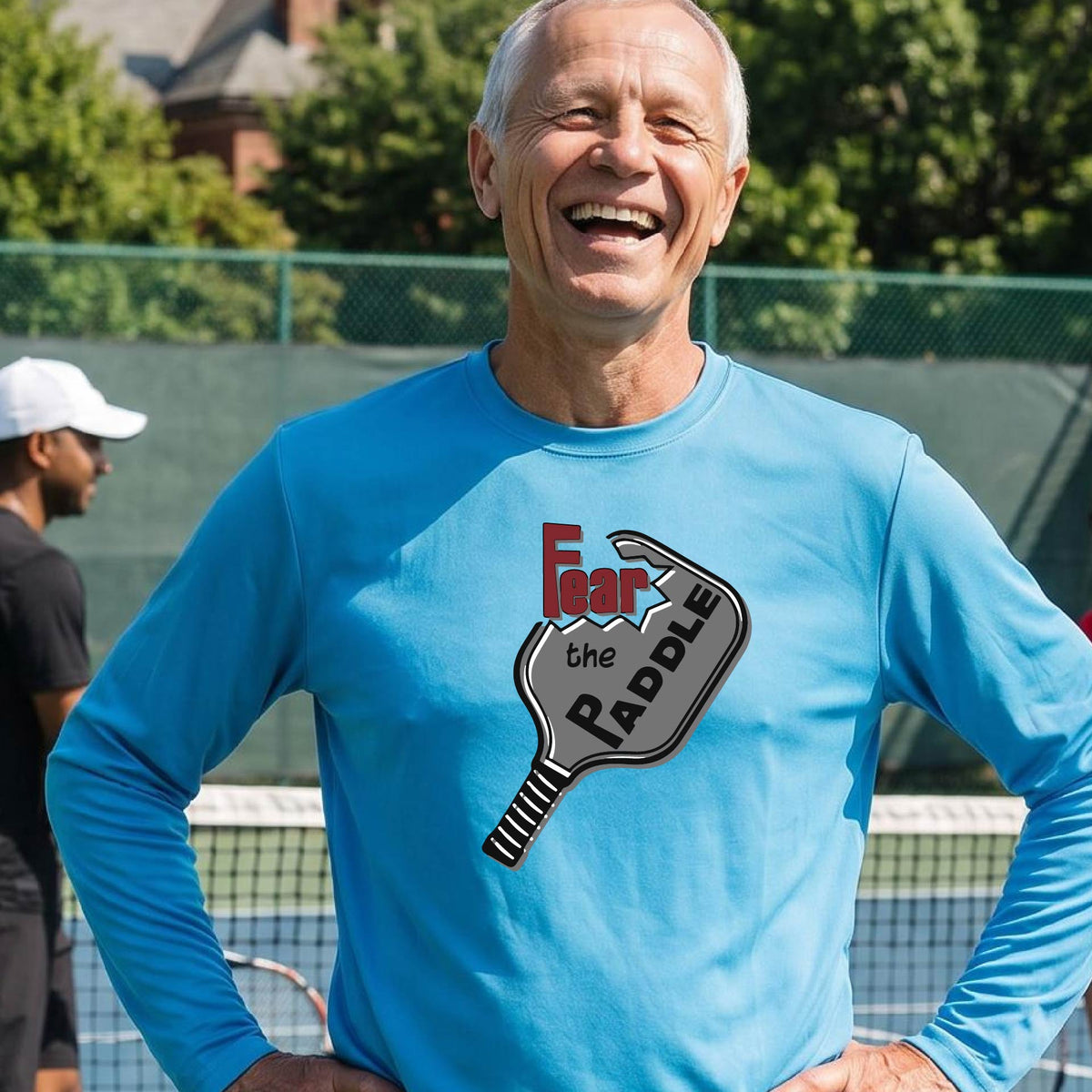 A man wearing a long-sleeve bright blue shirt with a pickleball graphic of the words 'FEAR THE PADDLE' cut into a pickleball paddle. 