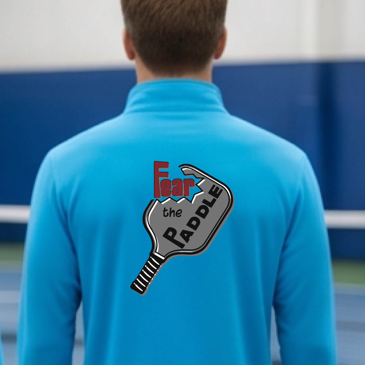 A man wearing a bright blue quarter-zip shirt with a pickleball graphic of the text 'FEAR THE PADDLE' cut into a pickleball paddle, standing on a pickleball court. 