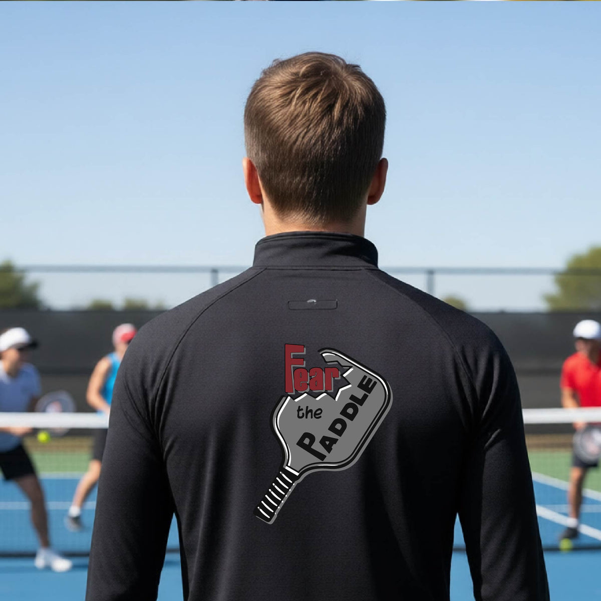 A man wearing a black quarter-zip shirt with a pickleball graphic of the text 'FEAR THE PADDLE' cut into a pickleball paddle, standing on a pickleball court. 