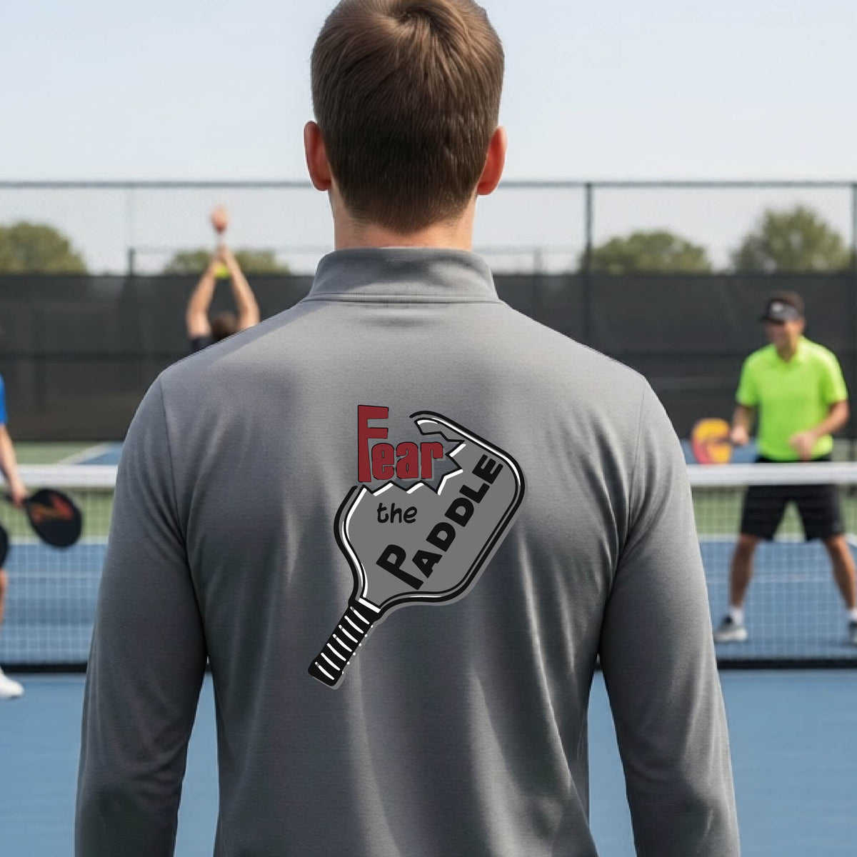 A man wearing a gray quarter-zip shirt with a pickleball graphic of the text 'FEAR THE PADDLE' cut into a pickleball paddle, standing on a pickleball court. 