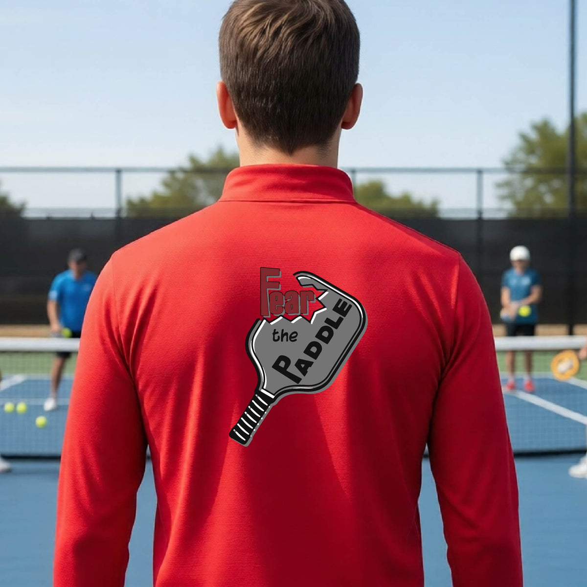 A man wearing a red quarter-zip shirt with a pickleball graphic of the text 'FEAR THE PADDLE' cut into a pickleball paddle, standing on a pickleball court. 