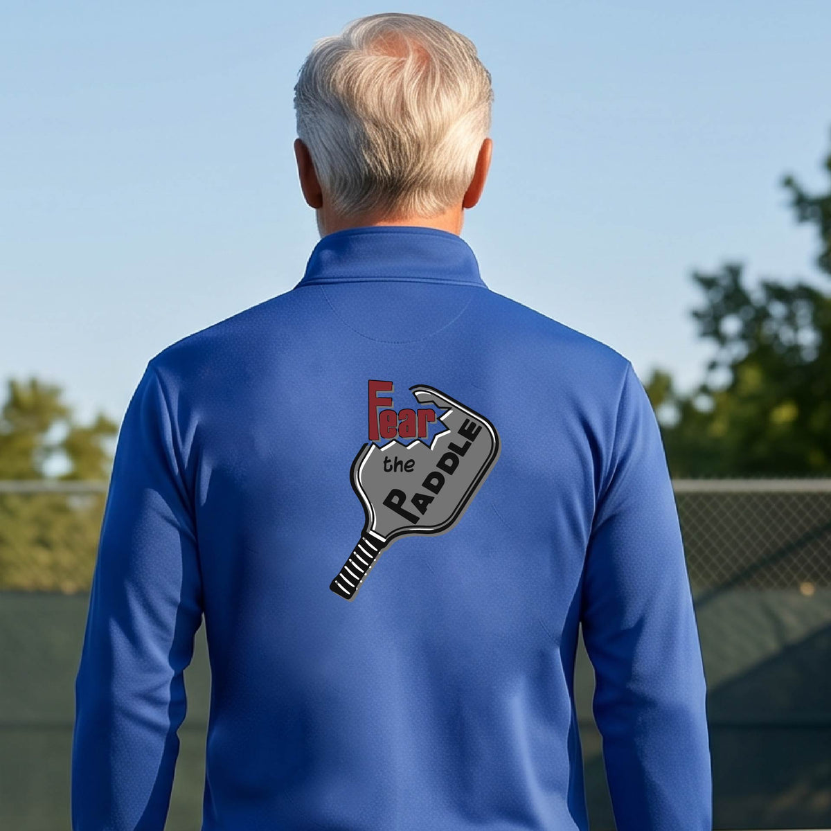 A man wearing a royal blue quarter-zip shirt with a pickleball graphic of the text 'FEAR THE PADDLE' cut into a pickleball paddle, standing on a pickleball court. 