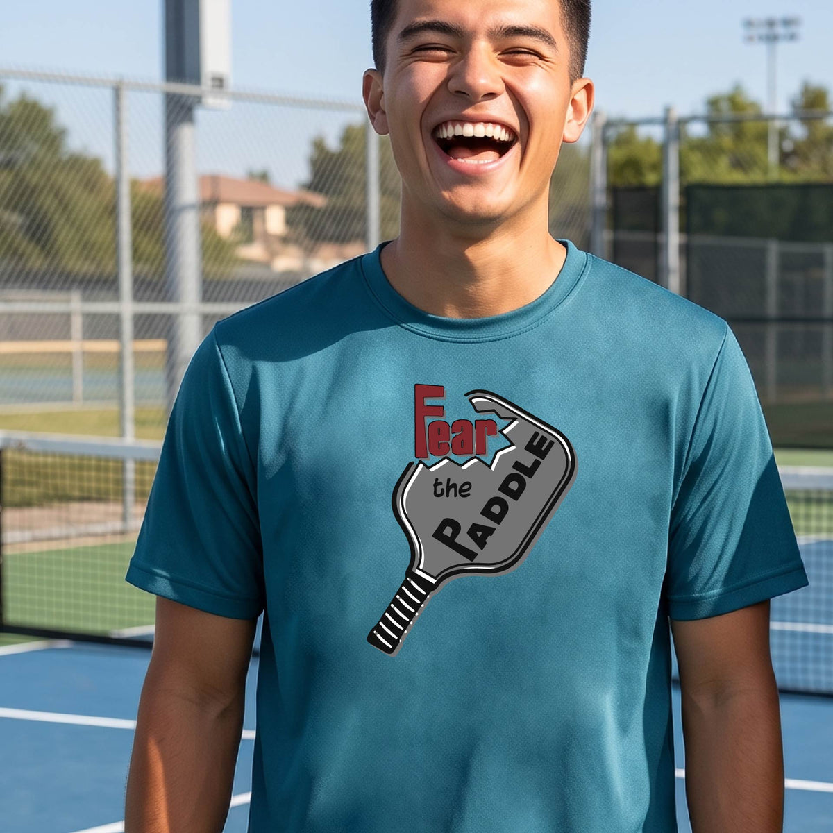A smiling man wearing a teal t-shirt with a graphic of a pickleball paddle and the words 'Fear the Paddle' in red and black, set against an outdoor background of a pickleball court. 