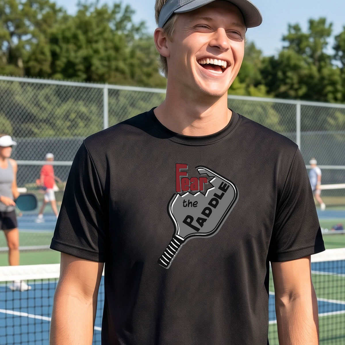 A smiling man wearing a black t-shirt with a graphic of a pickleball paddle and the words 'Fear the Paddle' in red and black, set against an outdoor background of a pickleball court. 