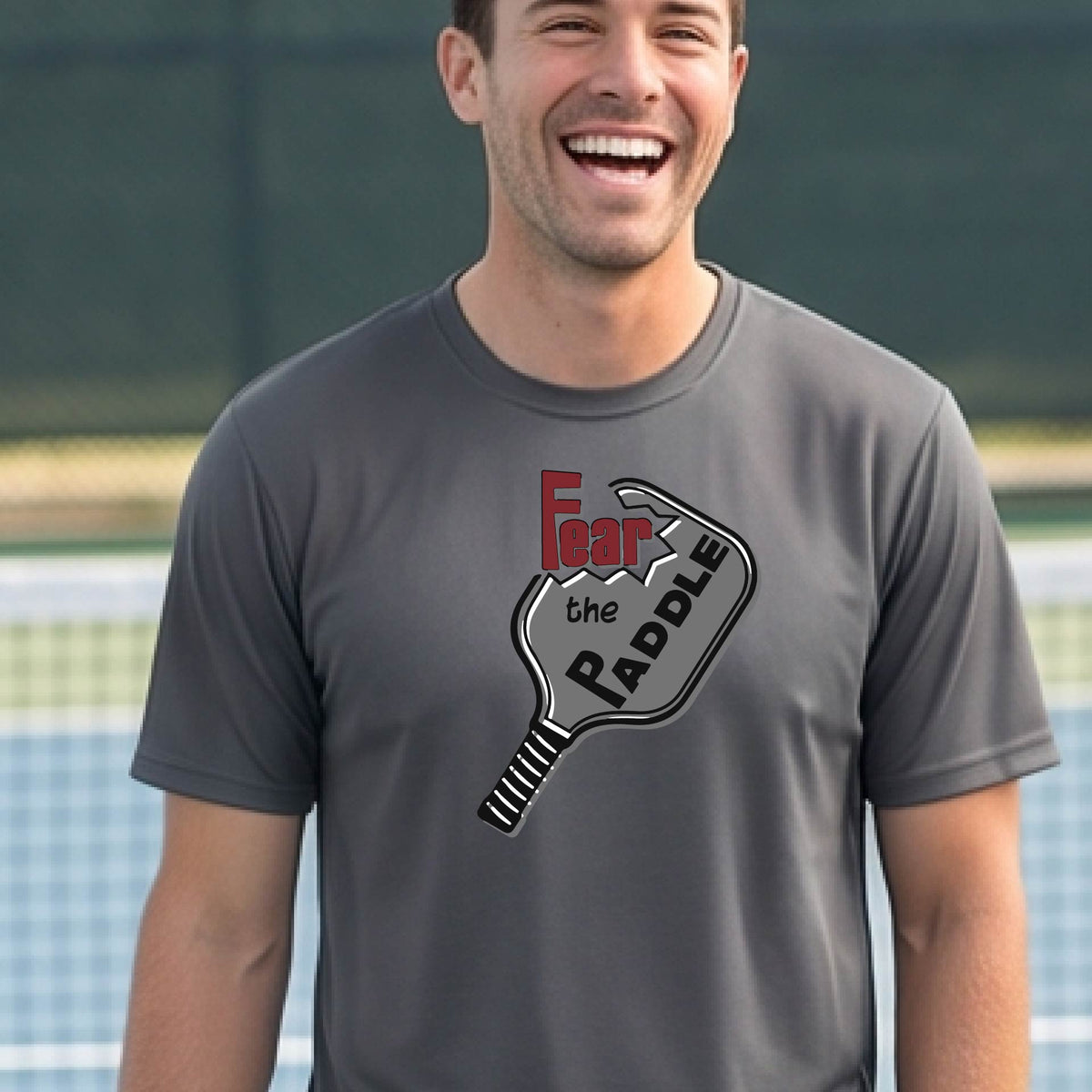 A smiling man wearing a gray t-shirt with a graphic of a pickleball paddle and the words 'Fear the Paddle' in red and black, set against an outdoor background of a pickleball court. 