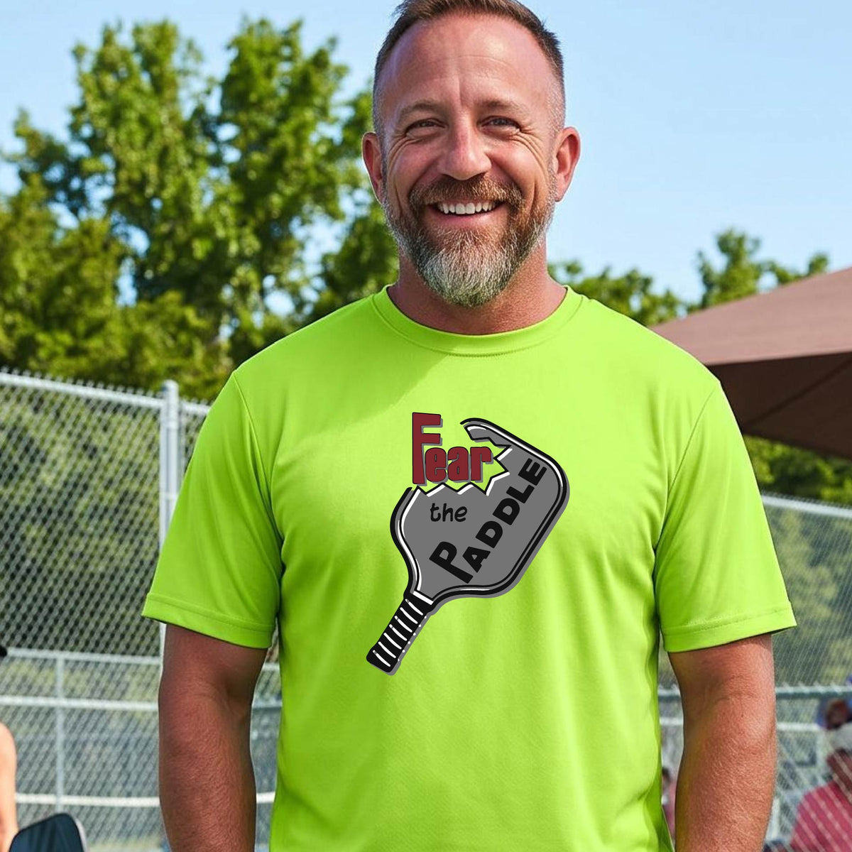 A smiling man wearing a bright neon yellow-green t-shirt with a graphic of a pickleball paddle and the words 'Fear the Paddle' in red and black, set against an outdoor background of a pickleball court. 
