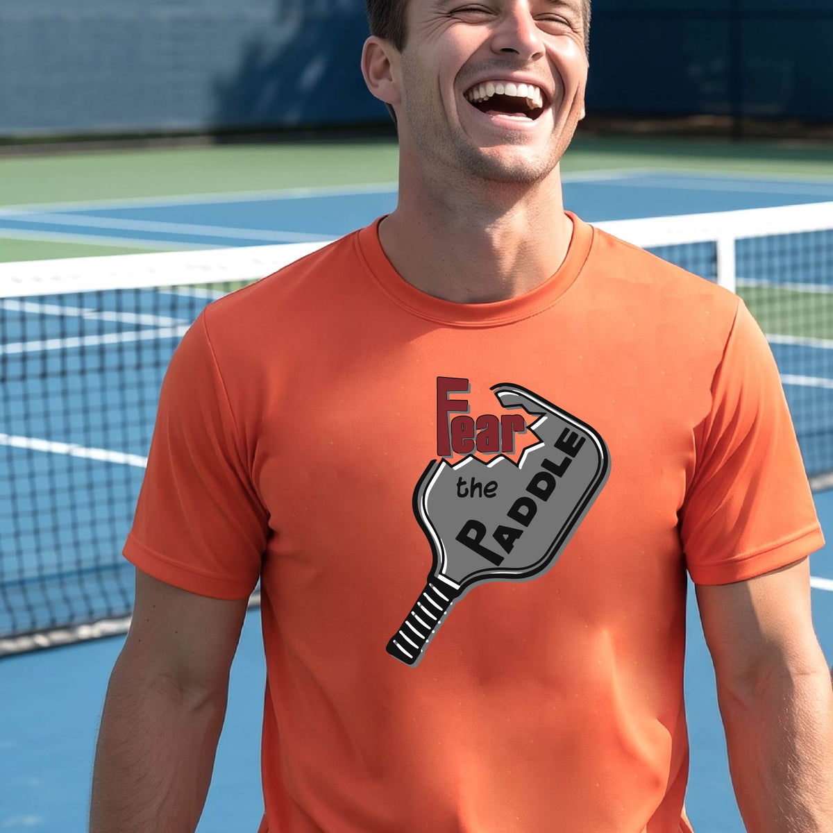A smiling man wearing an orange  t-shirt with a graphic of a pickleball paddle and the words 'Fear the Paddle' in red and black, set against an outdoor background of a pickleball court. 
