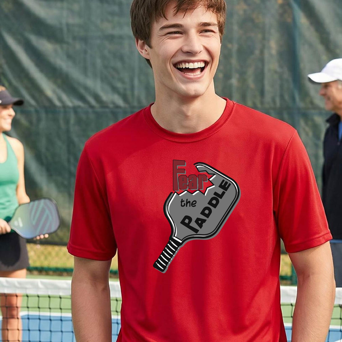 A smiling man wearing a red t-shirt with a graphic of a pickleball paddle and the words 'Fear the Paddle' in red and black, set against an outdoor background of a pickleball court. 