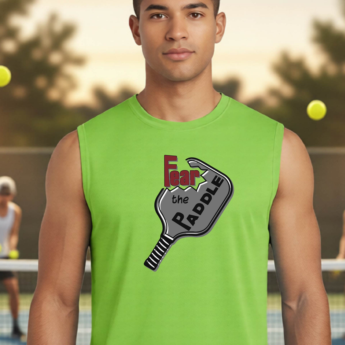 A man wearing a bright lime green sleeveless t-shirt with a graphic on the chest that reads 'Fear the PADDLE' in red and black over a paddle illustration. The background shows a blurred pickleball court