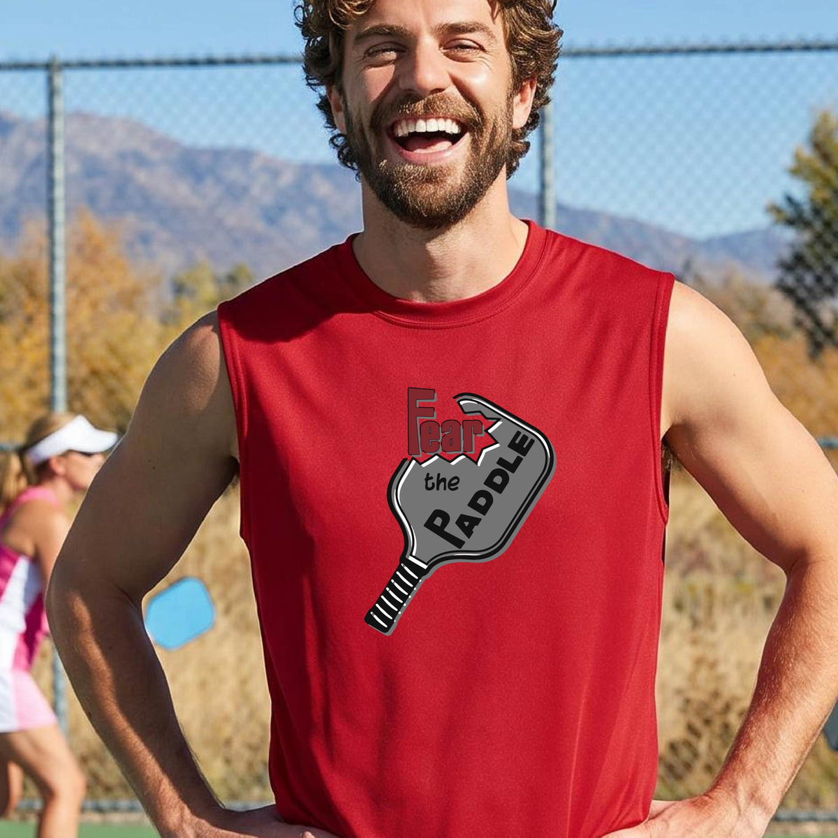 A man wearing a red sleeveless t-shirt with a graphic on the chest that reads 'Fear the PADDLE' in red and black over a paddle illustration. The background shows a blurred pickleball court