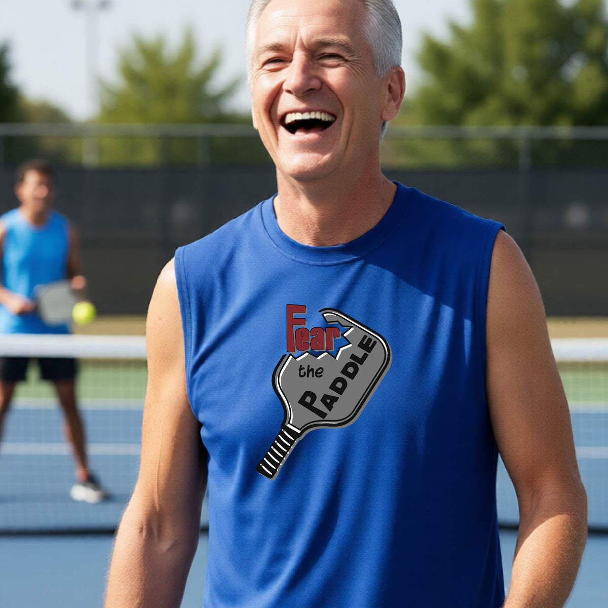 A man wearing a royal blue sleeveless t-shirt with a graphic on the chest that reads 'Fear the PADDLE' in red and black over a paddle illustration. The background shows a blurred pickleball court