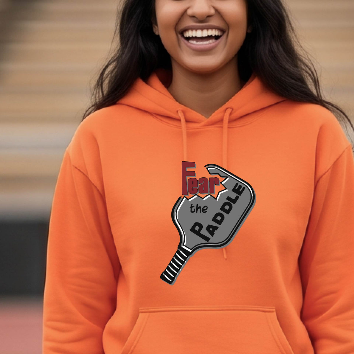 A woman wearing a bright orange hoodie pullover featuring a pickleball graphic of the text 'FEAR THE PADDLE' cut into a pickleball paddle, standing on a pickleball court. 