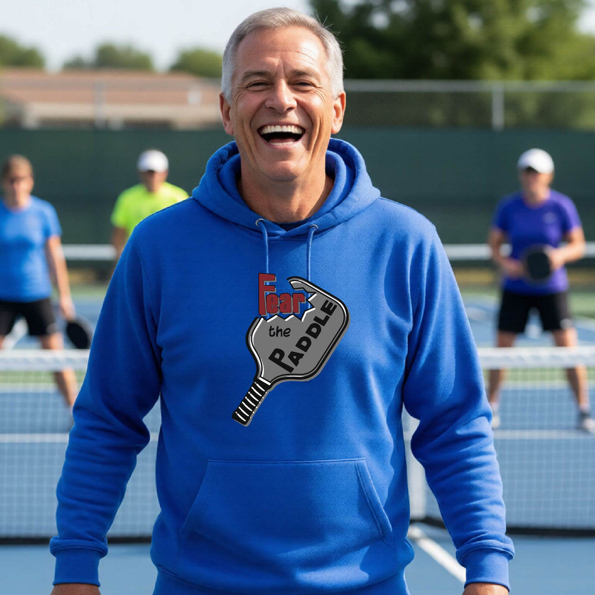 A man wearing a royal blue hoodie pullover featuring a pickleball graphic of the text 'FEAR THE PADDLE' cut into a pickleball paddle, standing on a pickleball court. 