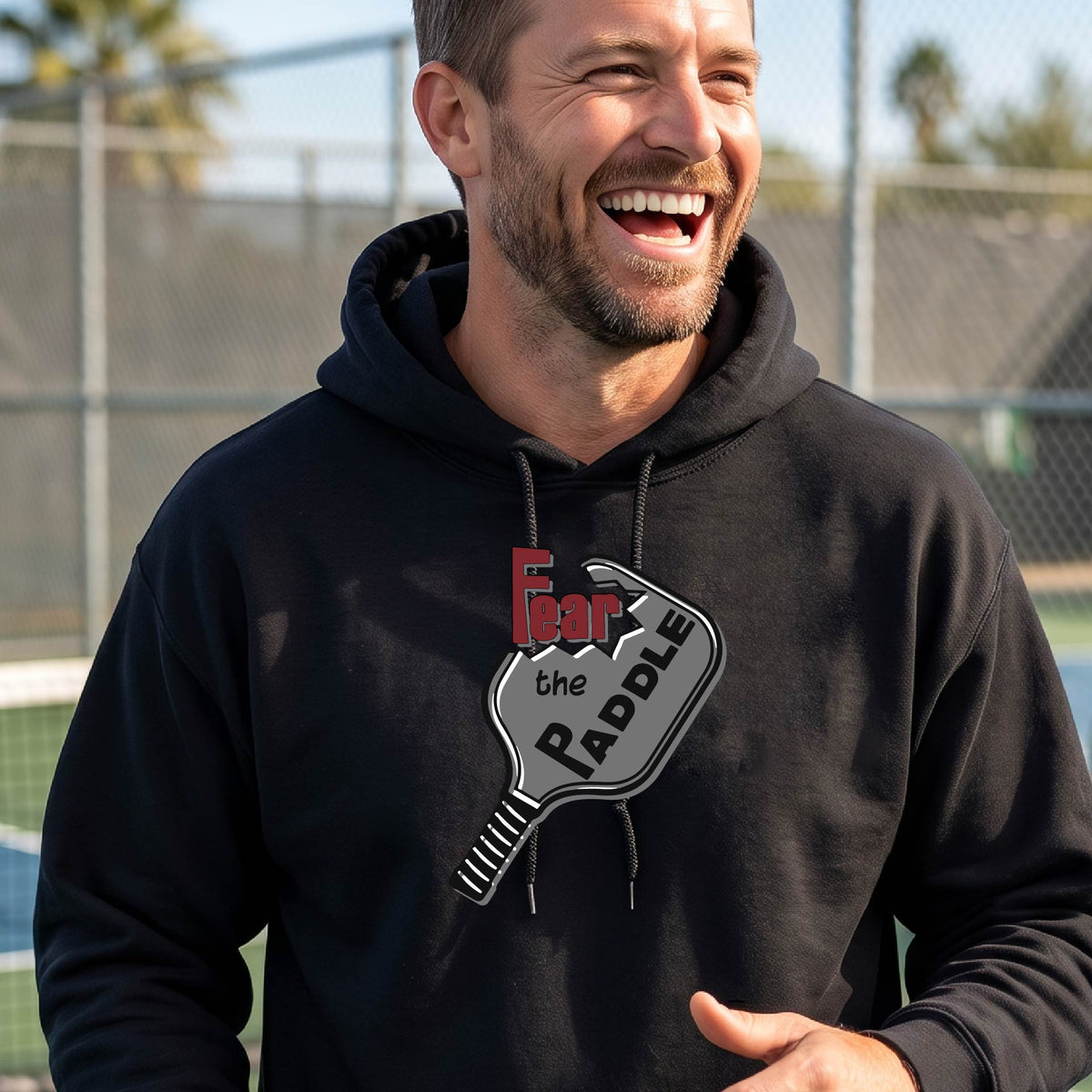 A man wearing a black hoodie pullover featuring a pickleball graphic of the text 'FEAR THE PADDLE' cut into a pickleball paddle, standing on a pickleball court. 